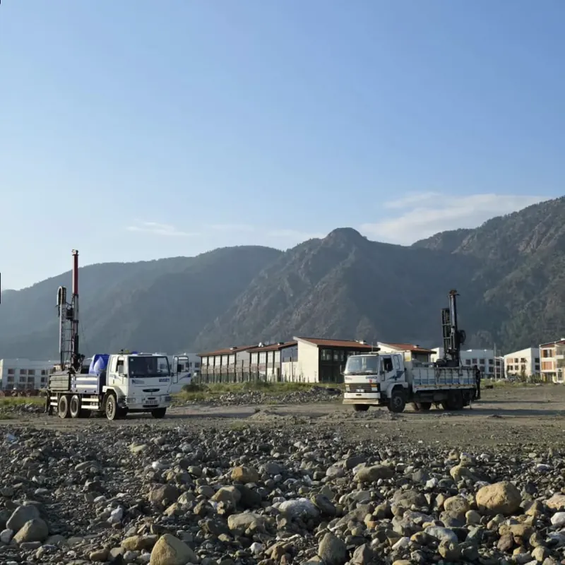 Two truck-mounted drilling rigs working simultaneously on a large site in front of mountains